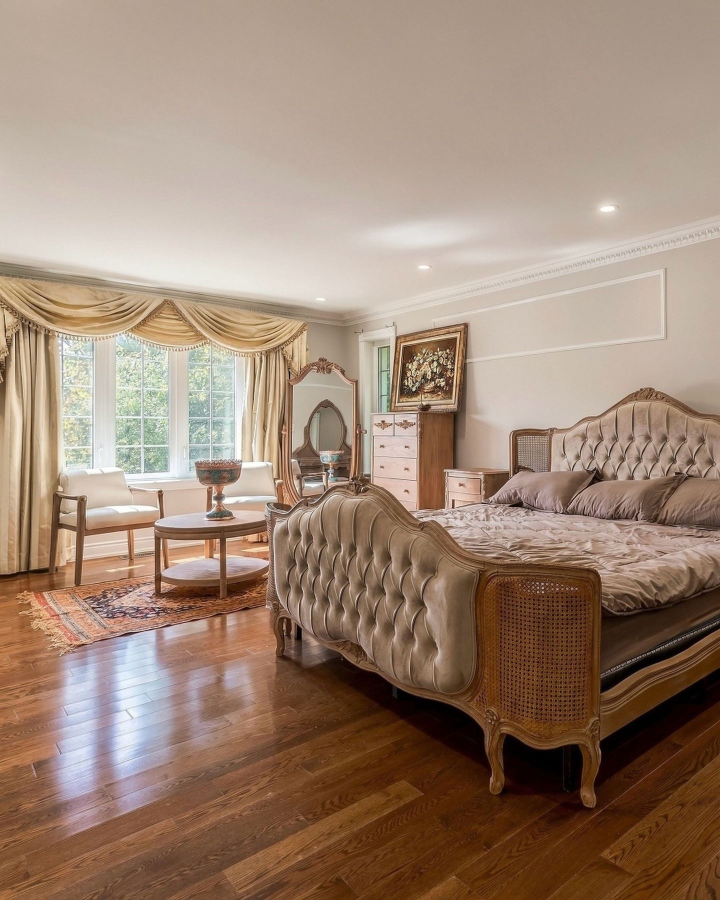 Sunlit primary bedroom with tufted bed, carved mirror and hardwood floors